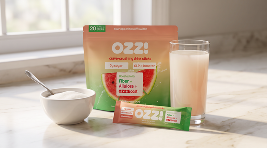 Bowl of white granulated sugar beside an Ozzi watermelon pouch, drink stick, and a glass of the prepared cloudy off-white drink on a marble kitchen counter