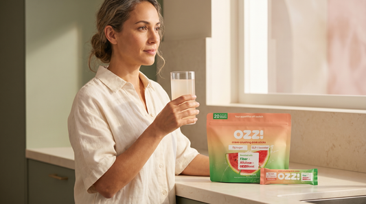 Woman holding a glass of the prepared Ozzi drink at a kitchen counter next to the OZZ! watermelon pouch and stick