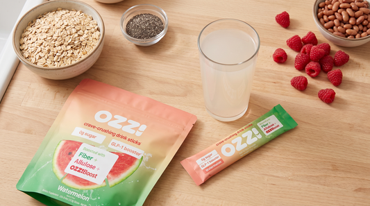 An overhead flatlay of an Ozzi watermelon pouch, a single stick, a prepared glass, and fiber-rich whole foods on a kitchen counter