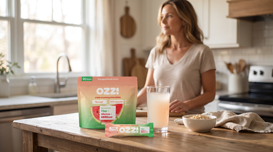 Woman at a sunlit kitchen island beside an Ozzi watermelon stick pack pouch, a single stick, and a glass of the prepared cloudy off-white drink