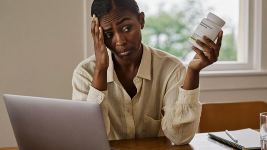 A woman in her late 30s at a kitchen table reading the back label of a supplement bottle with a skeptical expression