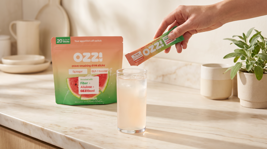 A hand holding an OZZ! watermelon drink stick above a glass of prepared Ozzi on a marble kitchen counter, with the OZZ! pouch behind it.