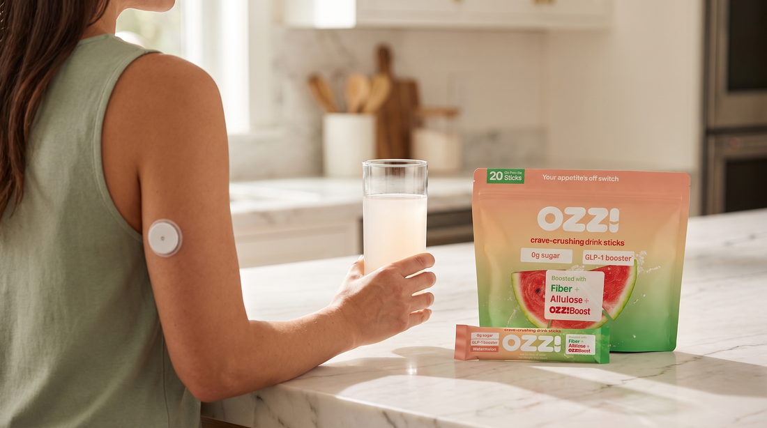 Woman with a continuous glucose monitor sensor on her arm holding a glass of the prepared Ozzi drink on a marble counter beside the Ozzi watermelon pouch and stick pack