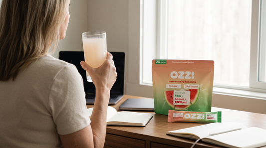 A woman at a home office desk holding a glass of prepared Ozzi, with the pouch and a stick pack on the desk