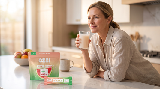 A woman in a bright kitchen holding a glass of prepared Ozzi watermelon drink beside the pouch and stick