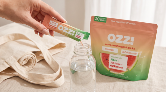A hand pouring an Ozzi watermelon drink stick into a water bottle beside the Ozzi pouch and a canvas tote bag