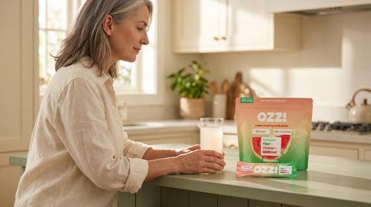 A woman in her early 50s preparing an Ozzi watermelon drink in a sunny kitchen, pouch and stick on the counter