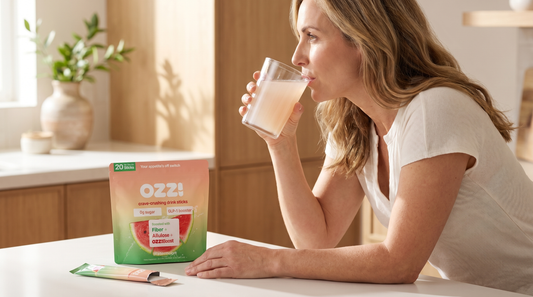Editorial photo of woman drinking the prepared Ozzi drink (cloudy off-white liquid with faint coral tint) at a sunlit kitchen counter with the OZZ! watermelon pouch beside her