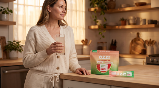 Woman in an evening kitchen calmly holding a glass of the prepared Ozzi watermelon drink, with the OZZ! pouch and stick pack on the counter