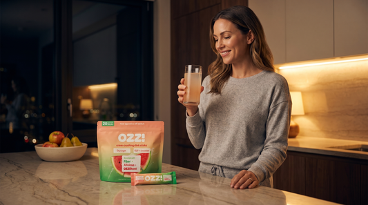 Woman in a kitchen at night holding a glass of prepared Ozzi watermelon drink, with the OZZ! pouch and a stick on the counter