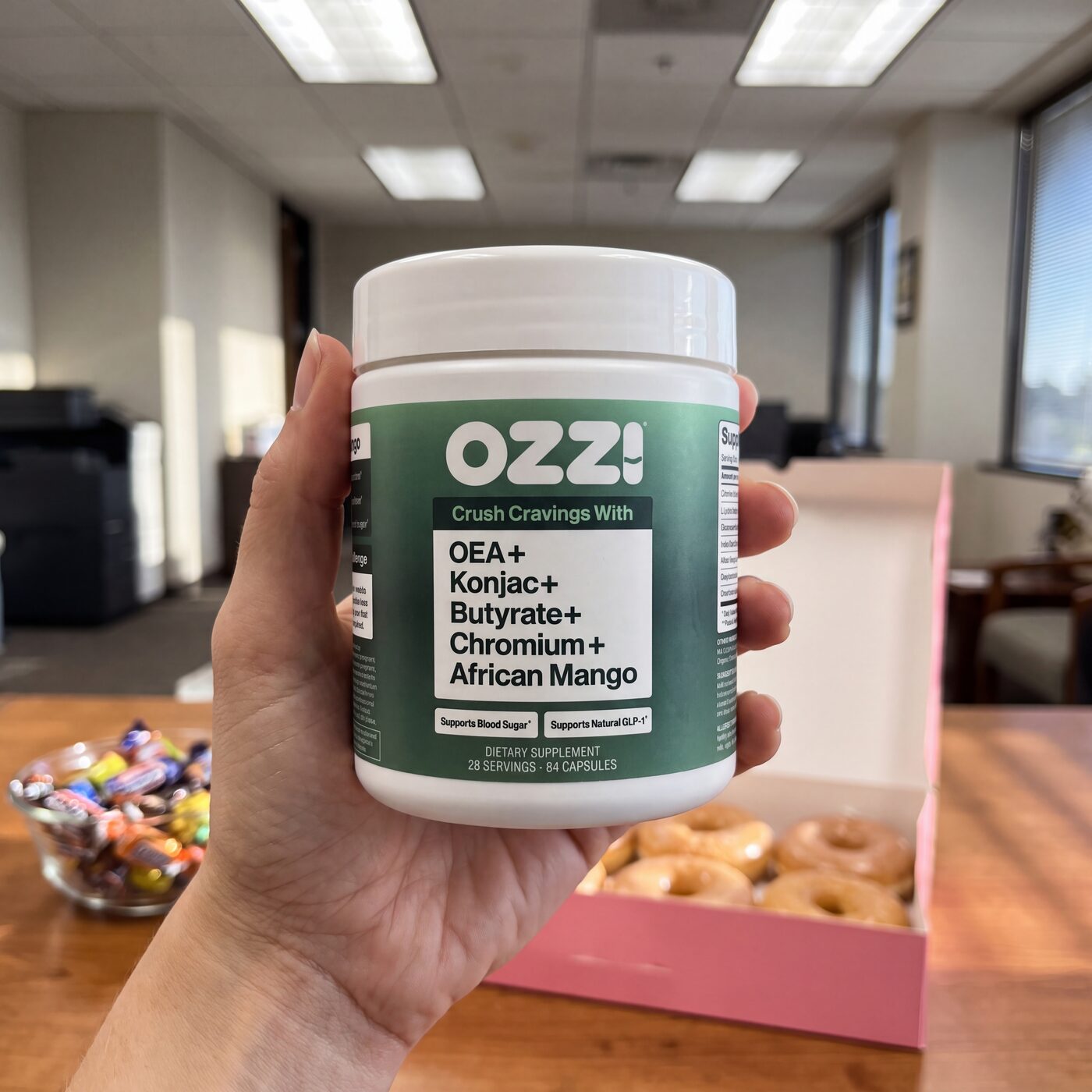 A hand holds a bottle of Ozzi natural GLP-1 supplement capsules in front of an office box of donuts and a bowl of candy