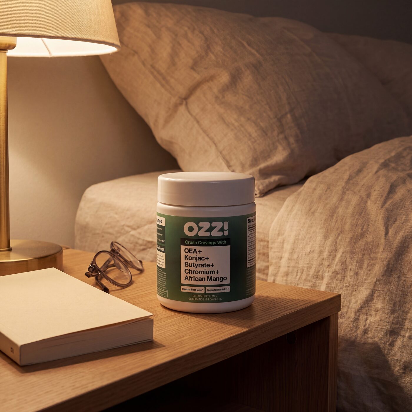 A bottle of Ozzi capsules on a nightstand beside a lamp, a book and reading glasses